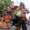 Two men in outfits made from lots of colourful ribbons playing a mobile drum set and a saxophone as they walk down a street procession