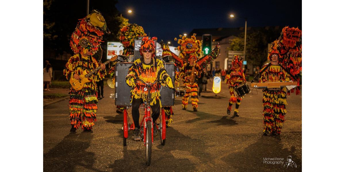 A band wearing outfits of brightly coloured ribbons walks down a street during a Torchlight Procession at night