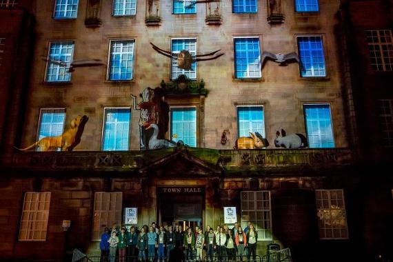 Rabbits, dogs, birds, and other creatures projected onto the front of Preston Town Hall whilst choir of children sing by doorway.