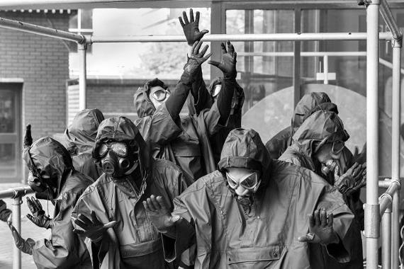 Arts group dressed in hazard clothing with gas masks, inside a box made from metal bars.