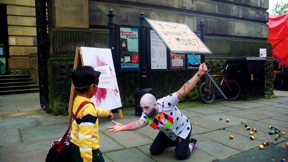 Street performer having a young boy help him pick up juggling balls outside the Harris.