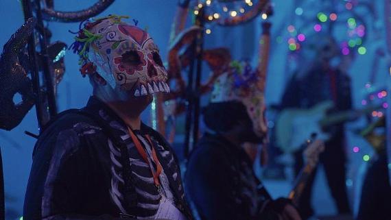 Band dressed with sugar skull masks performing music in dark tent lit by fairy lights.