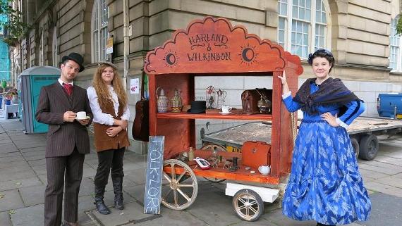 Two women and a man dressed in vintage clothing and stood by a cart with vintage memorabilia on display.
